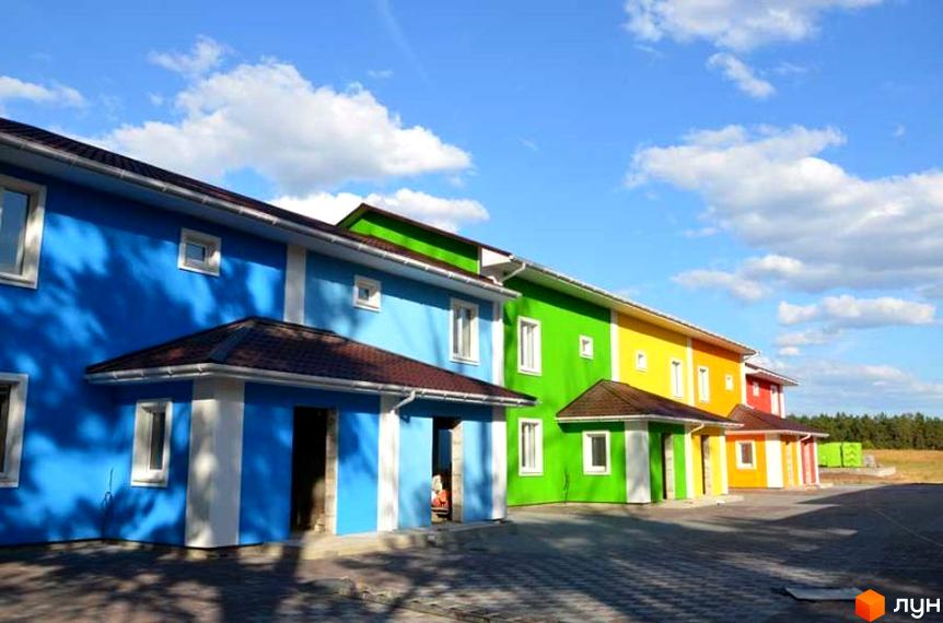 Яскраві таунхауси Ivankiv-townhouse у синьому, зеленому та жовтому кольорах під блакитним небом з хмарами. Прибудинкова територія з бруківкою та зеленими насадженнями.