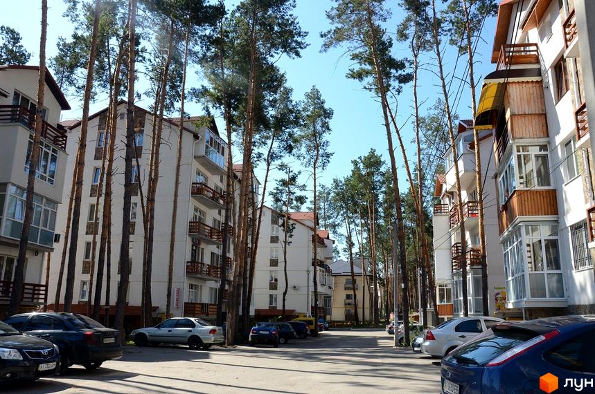 Житловий комплекс Островський: білі багатоповерхові будинки з дерев'яними балконами та соснами навколо. Парковка з автомобілями перед будинками.