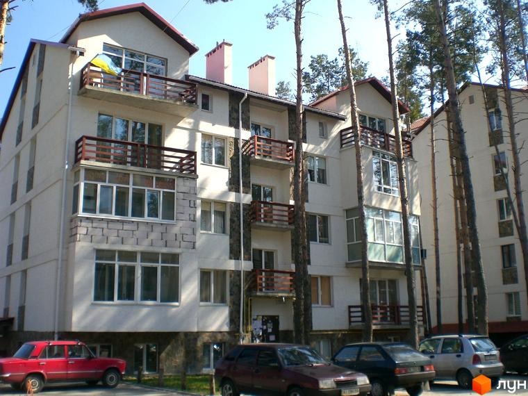 Зовнішній вигляд житлового комплексу Островський з балконами та вікнами. Навколо будинку ростуть високі сосни, а біля нього припарковані автомобілі.