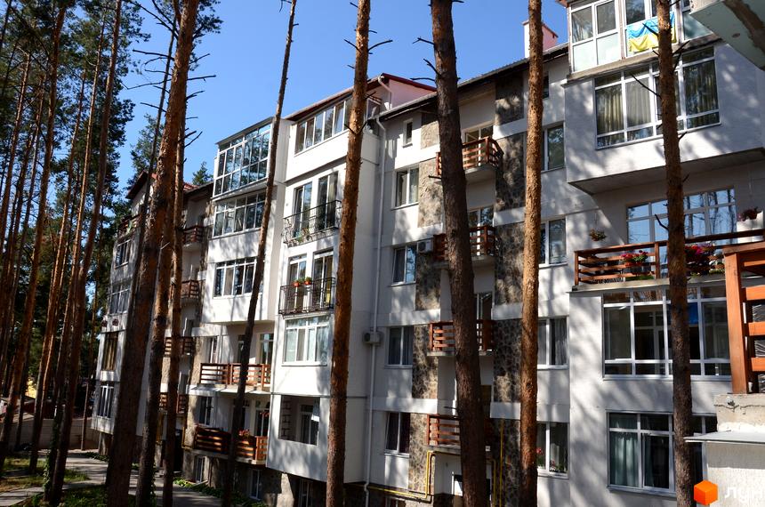 Житловий комплекс Островський: фасади будівель з балконами та великими вікнами, оточені високими соснами. Навколо новобудови зелена територія та доріжки.