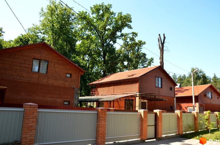 Будинок з дерев'яним фасадом та цегляною основою під червоним черепичним дахом. Огороджена територія з парканом та зеленими насадженнями.