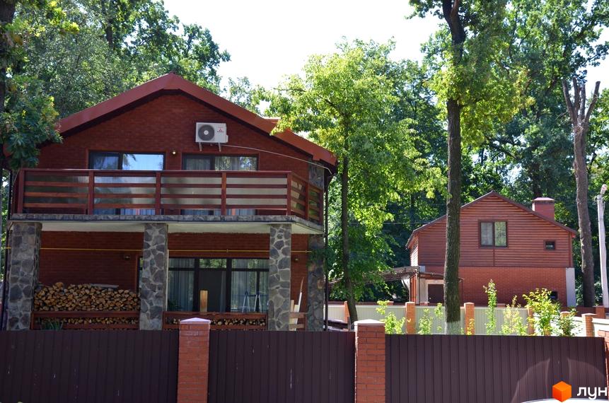 Двоповерховий цегляний будинок з балконом та дерев'яним парканом. Навколо будинку зелені дерева та кущі. Територія огороджена парканом.