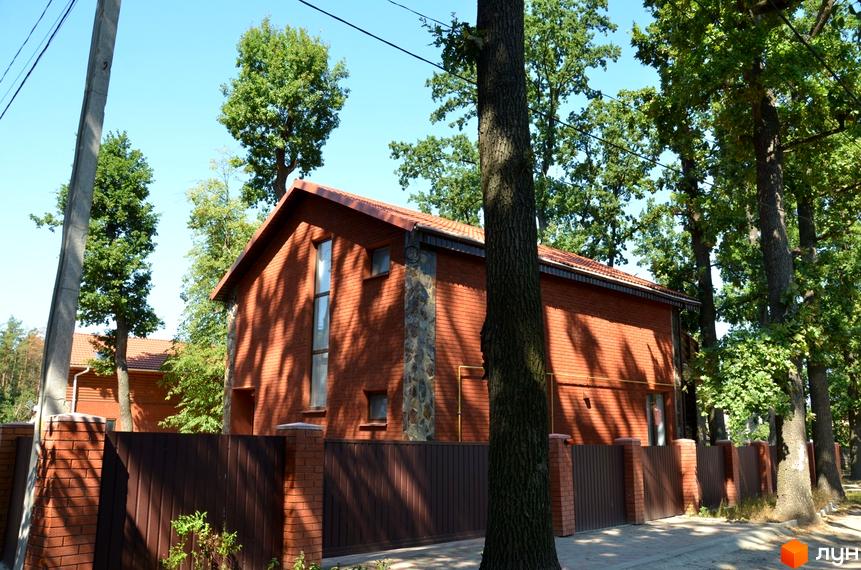 Будинок з червоної цегли з кам'яним оздобленням фасаду та коричневим парканом. Навколо будинку зелені дерева та блакитне небо.