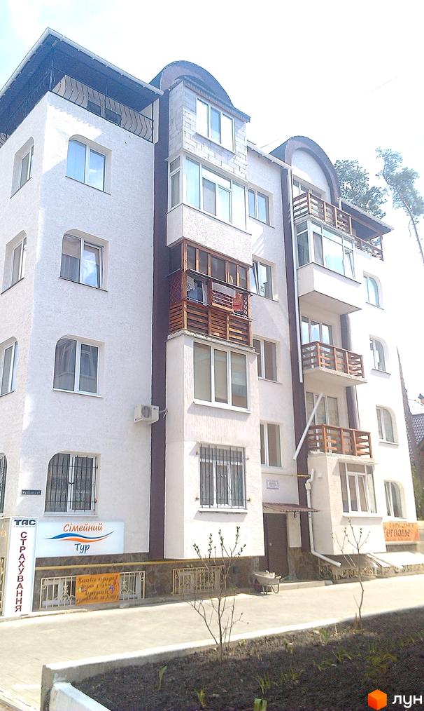 Зовнішній вигляд білої новобудови з дерев'яними балконами та вікнами. Прибудинкова територія з зеленими насадженнями та тротуаром.