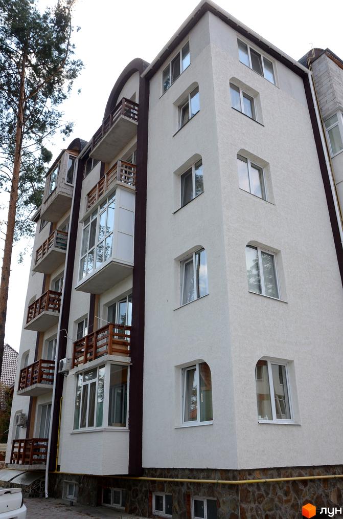 Зовнішній вигляд новобудови ЖК Затишний на вул. Університетській. Будинок має білий фасад, балкони з дерев'яними перилами та великі вікна. Навколо будинку видно дерева та елементи міського ландшафту.