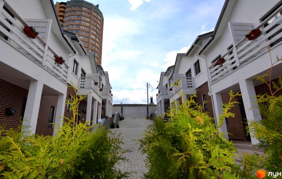 Таунхауси Сервус з білими фасадами та цегляними елементами, розташовані вздовж доглянутої алеї. На фоні видніється висотна будівля.