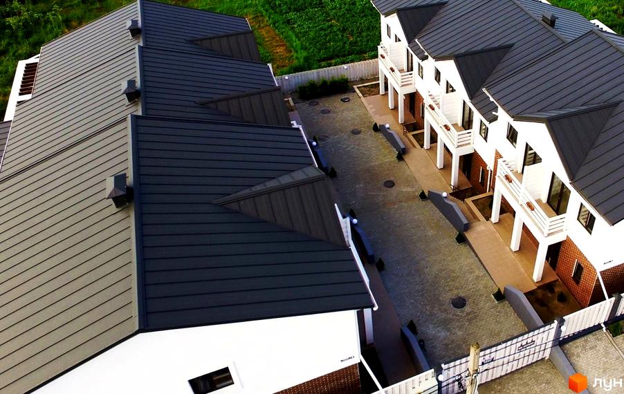 Зовнішній вигляд таунхаусів Сервус з темними металевими дахами та білими фасадами. Навколо будівель брукована територія та зелені насадження.