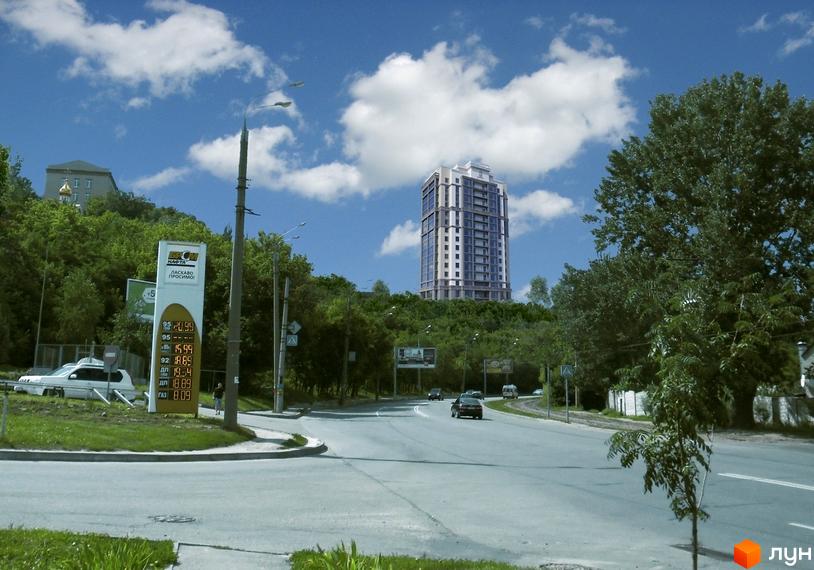 Новобудова ЖК Чумацький шлях на фоні зелених пагорбів та блакитного неба з хмарами. На передньому плані дорога з автомобілями та заправна станція.