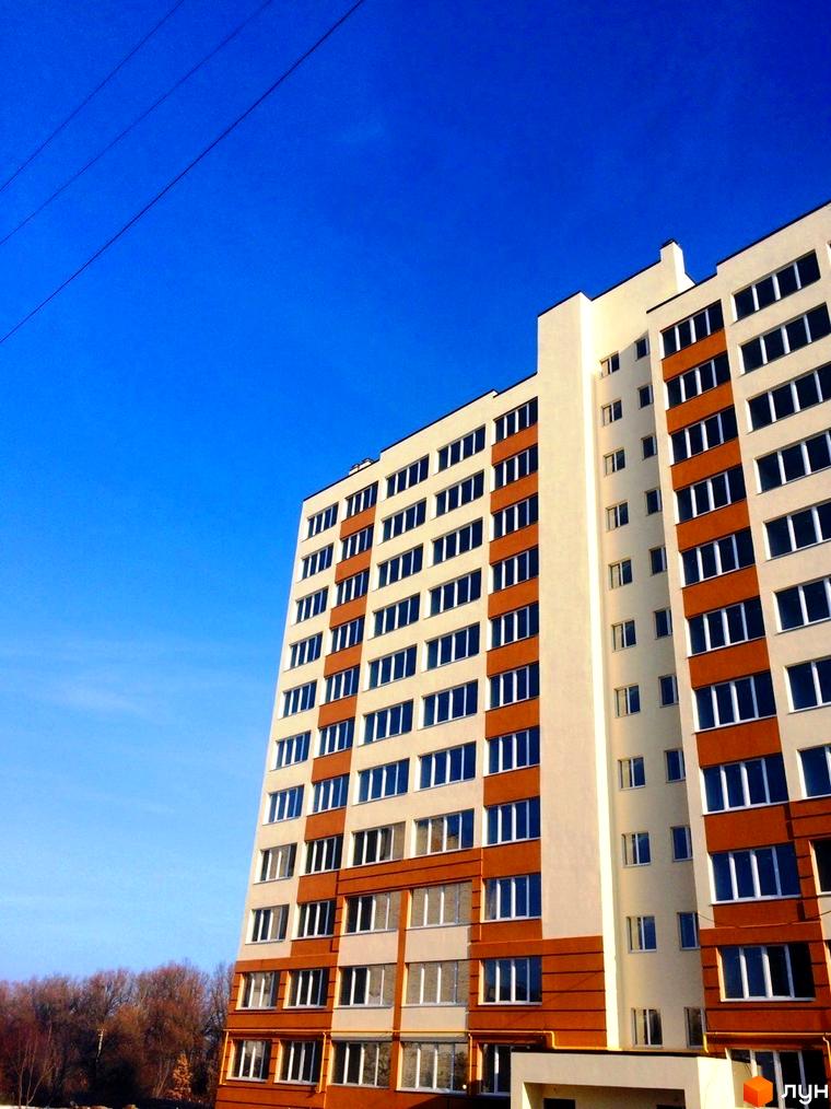 Зовнішній вигляд новобудови ЖК Затишна Оселя з біло-коричневими фасадами та численними вікнами на тлі ясного синього неба. Поруч видно дерева.