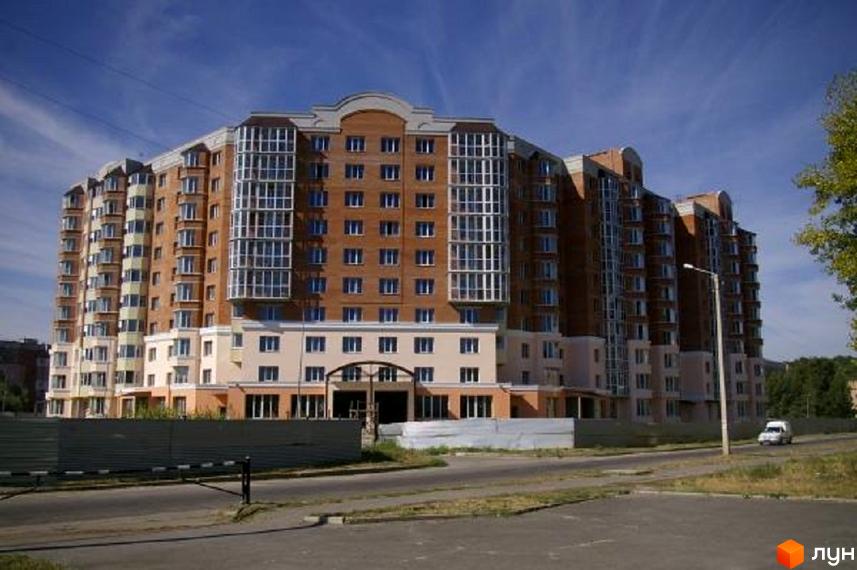 Висотна будівля житлового комплексу з цегляними фасадами та панорамними вікнами. На передньому плані асфальтована дорога та огорожа.