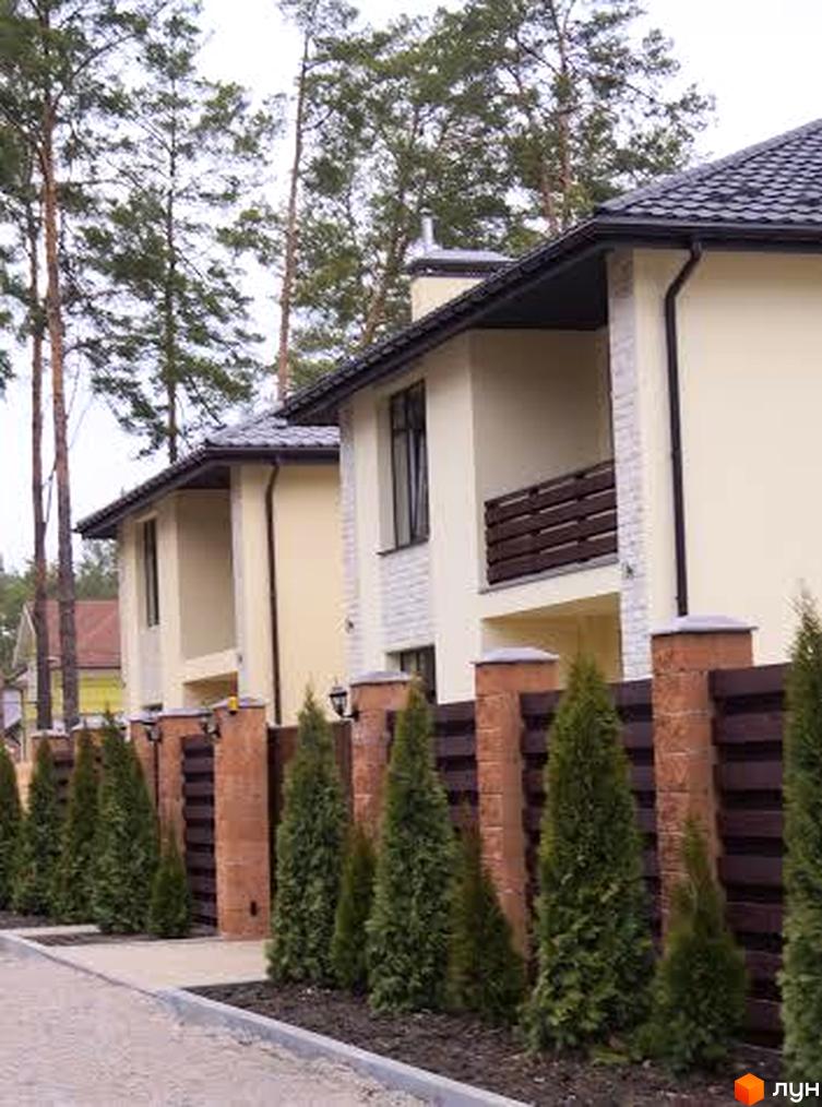 Зображення житлового будинку з двома поверхами з балконом та парканом з рослинами. На території комплексу висаджені туї.