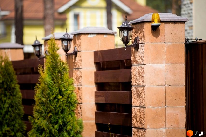 Огорожа Котеджного містечка Твій Дім з цегляними стовпами, ліхтарями та деревами. Вид на житлові будинки на задньому плані.