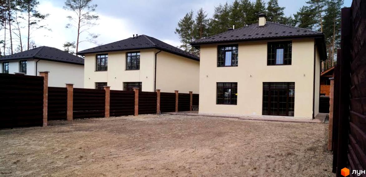 Зображення житлового будинку з двома поверхами та темним дахом. Перед будинком розташована прибудинкова територія з піщаним покриттям та огорожею.