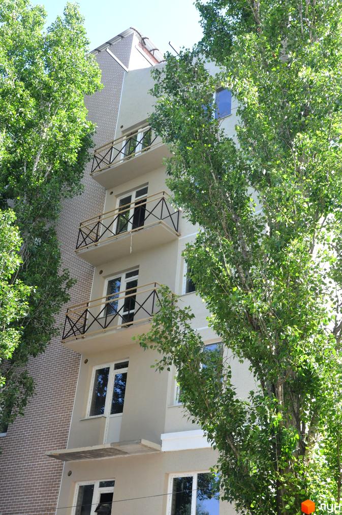 Багатоповерховий будинок з балконами та великими вікнами. Фасад будівлі світлого кольору, частково облицьований цеглою. Навколо будинку ростуть дерева.