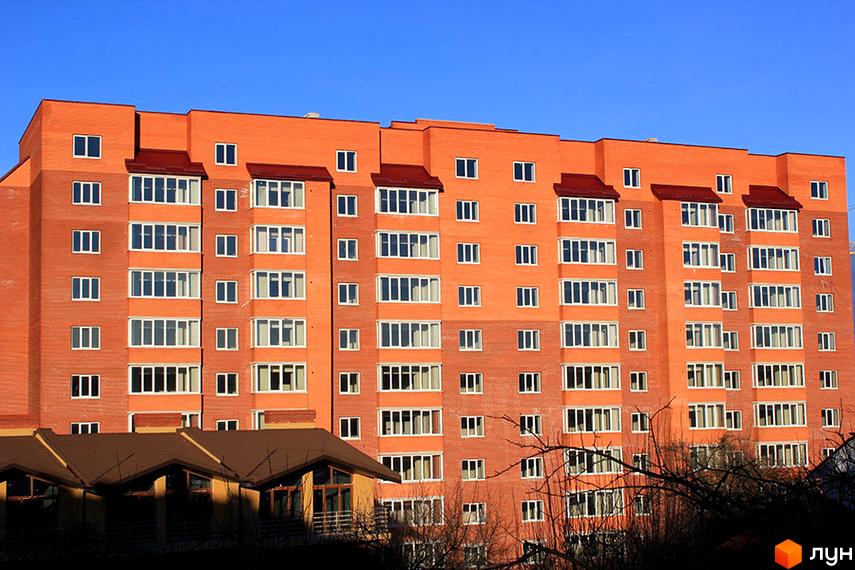 Висотна будівля житлового комплексу з цегляним фасадом та великою кількістю вікон. На передньому плані видно дерева та будівлі з коричневими дахами.