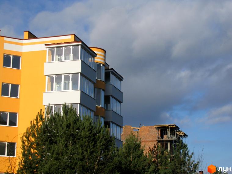 Житловий комплекс Приозерний. Фасад будинку з жовтими та білими елементами, заскленими балконами. Навколо зелені дерева та хмарне небо.