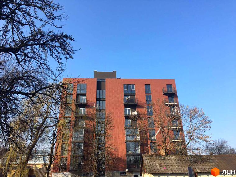 Висотна червона будівля ЖК на вулиці Дубравського, 5. Фасад з вікнами та балконами, навколо дерева та блакитне небо.