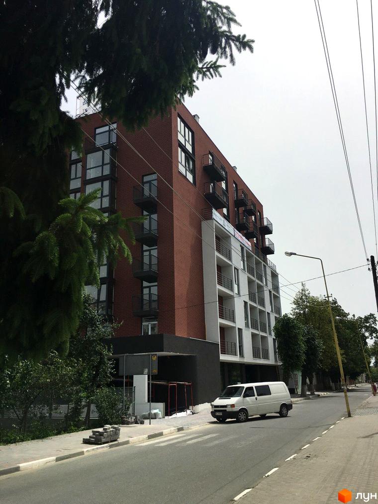 Зовнішній вигляд багатоповерхового будинку з цегляним фасадом та балконами. На прибудинковій території видно дорогу, тротуар та зелені насадження.