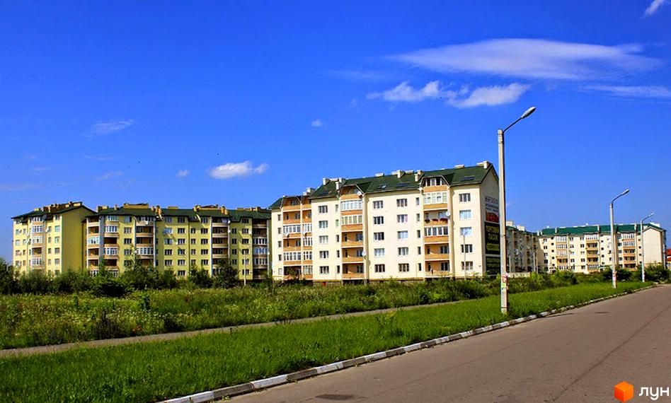 Зображення житлового комплексу з багатоповерховими будинками. На передньому плані зелена трава та дорога.