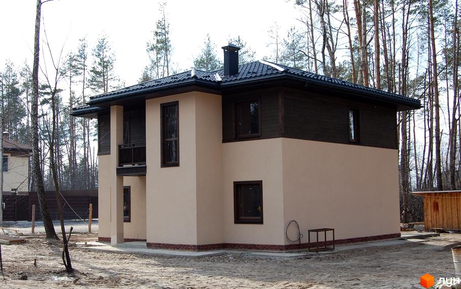 Котеджне містечко КМ Forest Family Avenue: двоповерховий будинок з бежевим фасадом, темним дахом та дерев'яними елементами. Навколо будинку піщана ділянка з деревами та парканом.