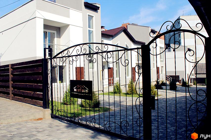 Фасад таунхаусів Таунхаус „Емірати“ з білими стінами та темними дахами, а також доглянута прибудинкова територія з бруківкою та зеленню.