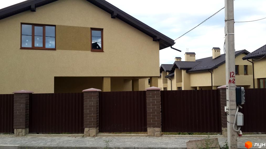 Будинки житлового комплексу з бежевими фасадами та коричневими дахами. Перед будинками розташований коричневий паркан з цегляними стовпами та тротуар.