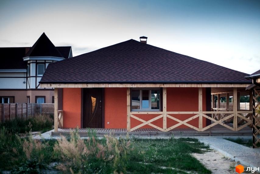 Одноповерховий будинок з помаранчевим фасадом, коричневим дахом та дерев'яною терасою в Котеджному містечку HappyTown. На передньому плані трава та доріжка.