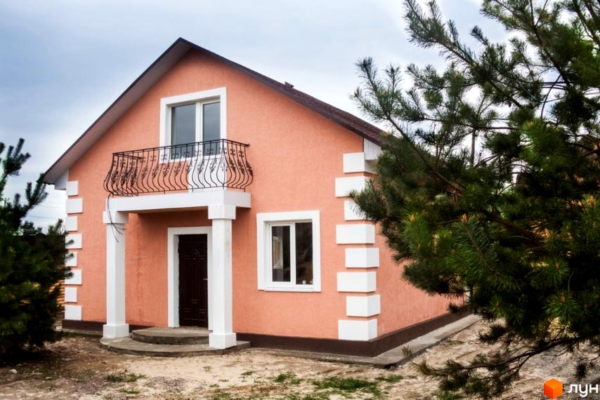 Будинок персикового кольору з темним дахом, білими вікнами та балконом в Котеджному містечку HappyTown. Прибудинкова територія з ґрунтом та хвойними деревами.