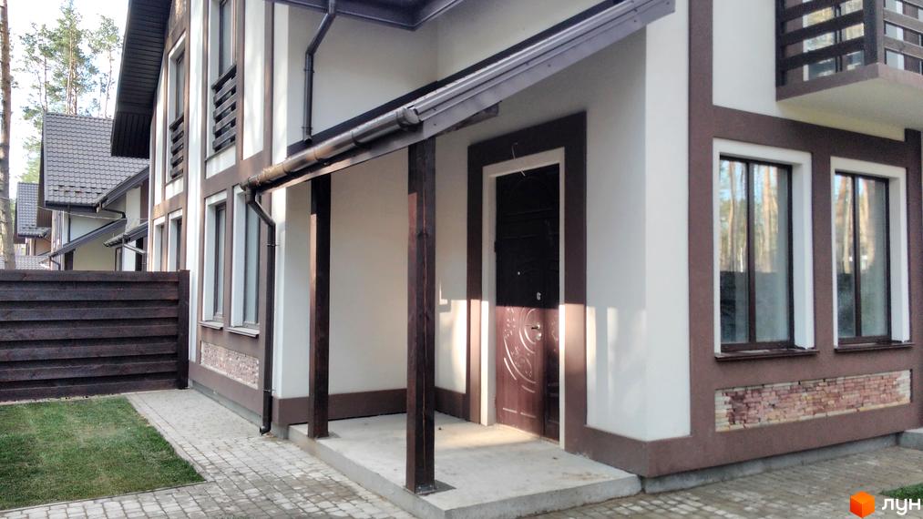 Двоповерховий будинок з білими стінами та коричневими елементами. Перед будинком є тераса з дерев'яними колонами, а також доріжка з бруківки та газон.