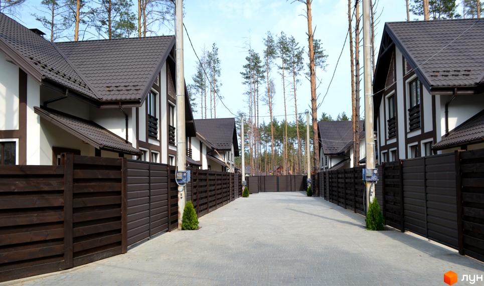 Зображення житлового комплексу КМ Едельвейс. Будинки з коричневими дахами та огорожами, розташовані вздовж вимощеної дороги. Навколо будівель ростуть дерева.