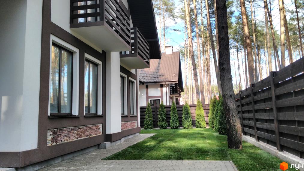 Зображення будинку в Котеджному містечку Едельвейс. Двоповерхова будівля з балконами, вікнами та цегляною кладкою біля основи, зелений газон та дерева.