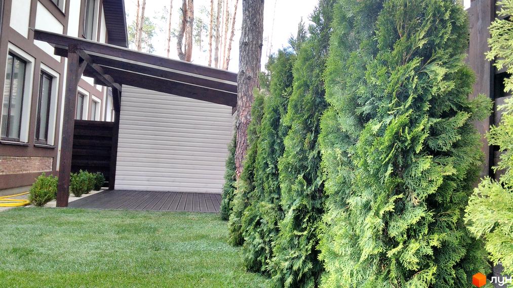 Зображення фасаду будинку з білими стінами та коричневим дахом. Перед будинком зелена трава та ряд хвойних дерев.