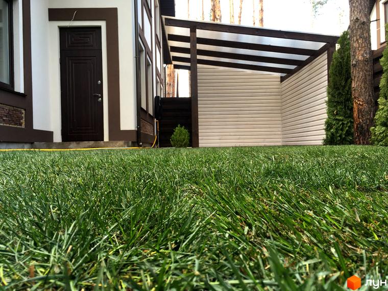 Зображення будинку з темними дверима та коричневими оздобленнями. Перед будинком зелена трава, а також видно навіс та дерева.
