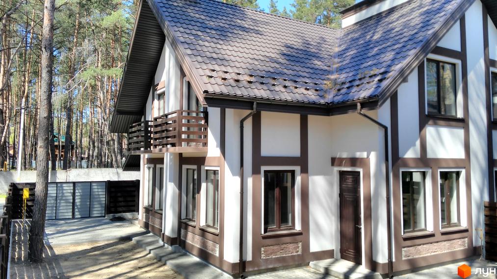 Зображення фасаду Котеджного містечка Едельвейс. Двоповерховий будинок з балконом, коричневим дахом та білими стінами, розташований серед соснового лісу.