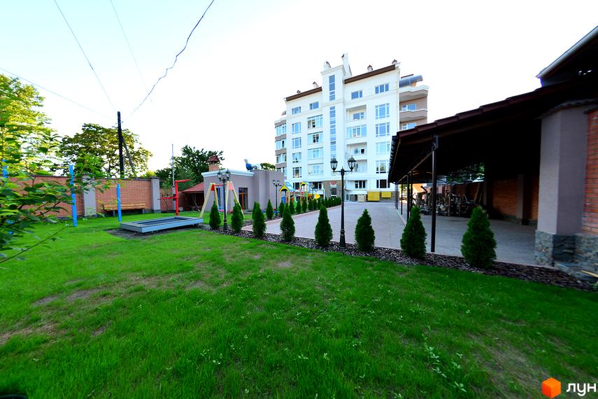 Багатоповерховий житловий будинок світлого кольору з великими вікнами. Перед ним зелений газон, доріжка з ліхтарями та дитячий майданчик.