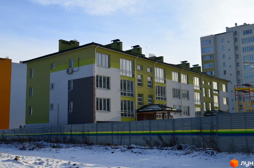 Зображення фасаду та прибудинкової території Новобудови Новий. Багатоповерховий будинок з великими вікнами та кольоровим фасадом на фоні зимового пейзажу.