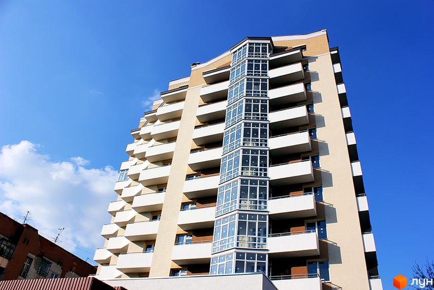 Зображення багатоповерхового будинку на вулиці Угорська, 7а. Житловий комплекс з великими балконами та панорамними вікнами на тлі блакитного неба.