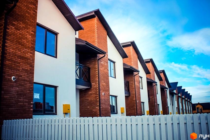 Котеджне містечко КМ Струмочок: фасади будинків з цегляними елементами та білою штукатуркою, балкони, вікна. Огороджена територія з білим парканом.