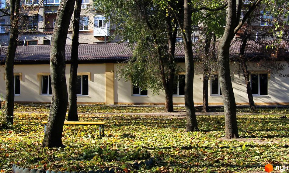 Одноповерхова будівля житлового комплексу Етюд зі світлими стінами та коричневим дахом, розташована серед дерев з осіннім листям.