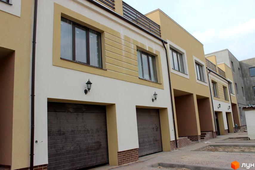 Зовнішній вигляд новобудови на вулиці Кибалюка, 17. Фасад будинку оздоблений світлими та бежевими кольорами, з великими вікнами та гаражними воротами. Прибудинкова територія облаштована тротуарною плиткою та зеленими насадженнями.