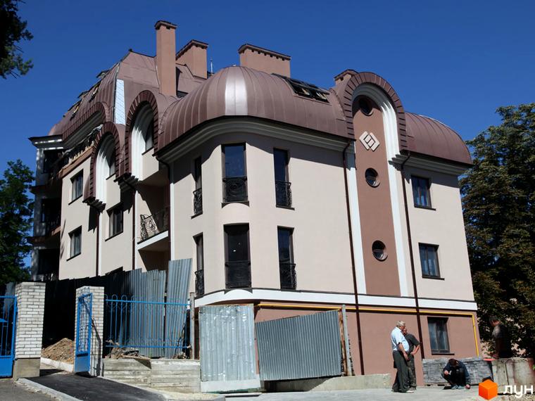 Зображення фасаду будинку за адресою: вул. Братів Тимошенків, 2б. Багатоповерхова будівля з коричневим дахом та світлим фасадом, видно частину прибудинкової території.