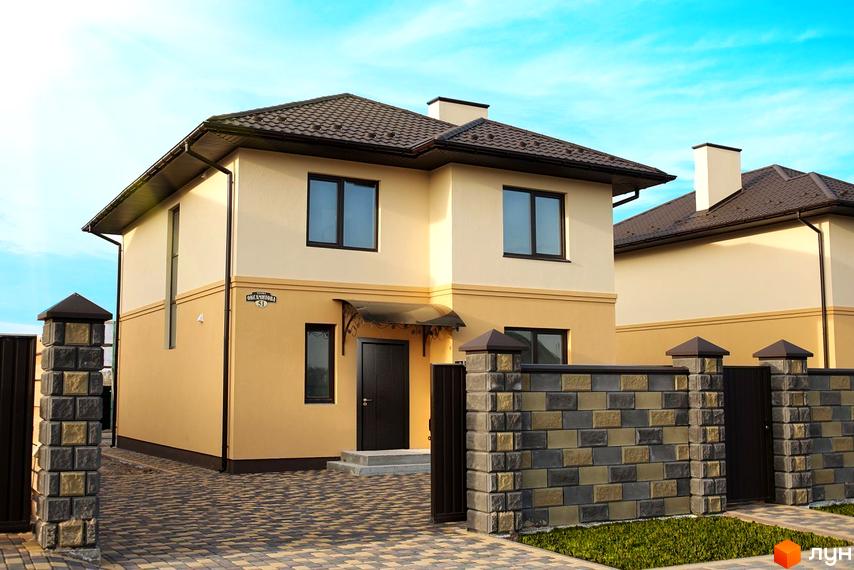 Двоповерховий будинок Котеджного містечка Оксамит з бежевим фасадом та коричневим дахом. Прибудинкова територія має брукований двір та цегляний паркан.