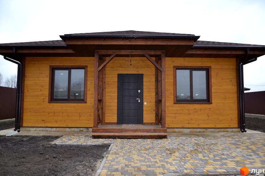 Одноповерховий будинок з дерев'яним фасадом та темним дахом. Перед будинком викладена бруківка, що створює акуратний вигляд.