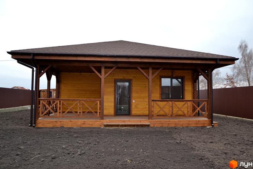 Одноповерховий дерев'яний будинок з терасою та коричневим дахом. Навколо будинку земля, огороджена коричневим парканом.