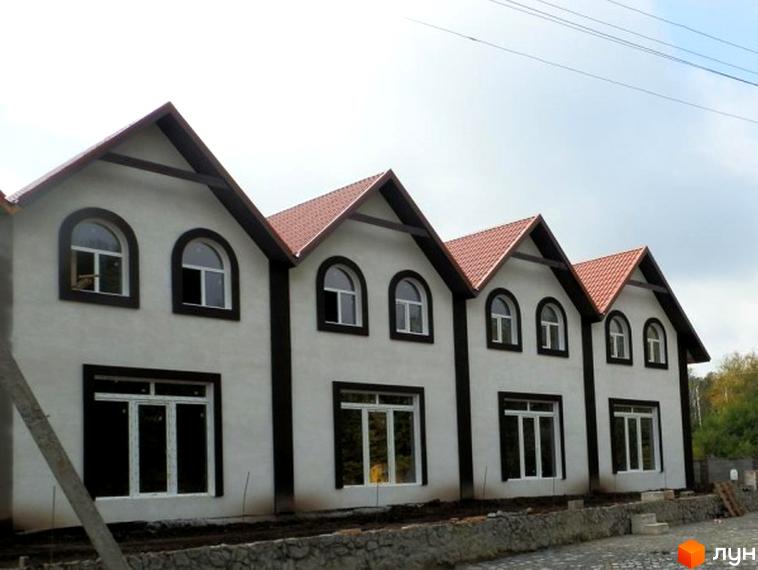 Зображення житлового комплексу таунхаусів з білими фасадами, темними віконними рамами та червоними дахами. На передньому плані видно прибудинкову територію з кам'яною кладкою.