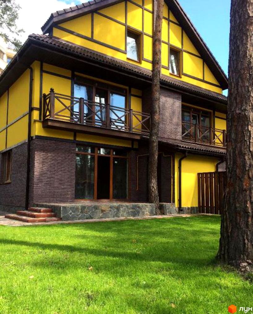 Котеджне містечко Кентербері: жовтий будинок з коричневим каркасом та цегляною основою, дерев'яними балконами. Навколо зелений газон та дерева.