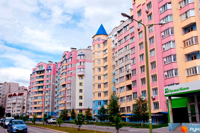 Зовнішній вигляд житлового комплексу Мікрорайон „Поділля“ з різнокольоровими фасадами та блакитним дахом. Поруч дорога з автомобілями та зелені насадження.