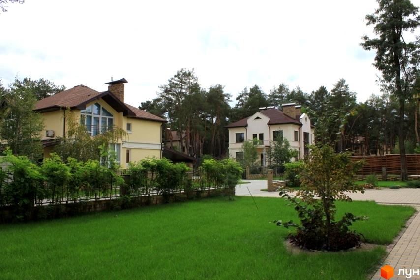 Двоповерхові будинки з коричневою черепицею та світлими стінами. На території Котеджне містечко Шишкино є газон, дерева та доріжки з бруківки.