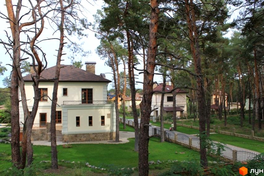 Білий будинок з коричневим дахом серед дерев. Котеджне містечко Шишкино з доглянутою територією та зеленими газонами.