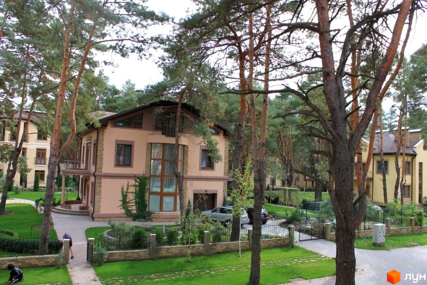 Двоповерховий будинок з великими вікнами та балконом. На території Котеджне містечко Шишкино є доглянутий газон, доріжки та дерева.